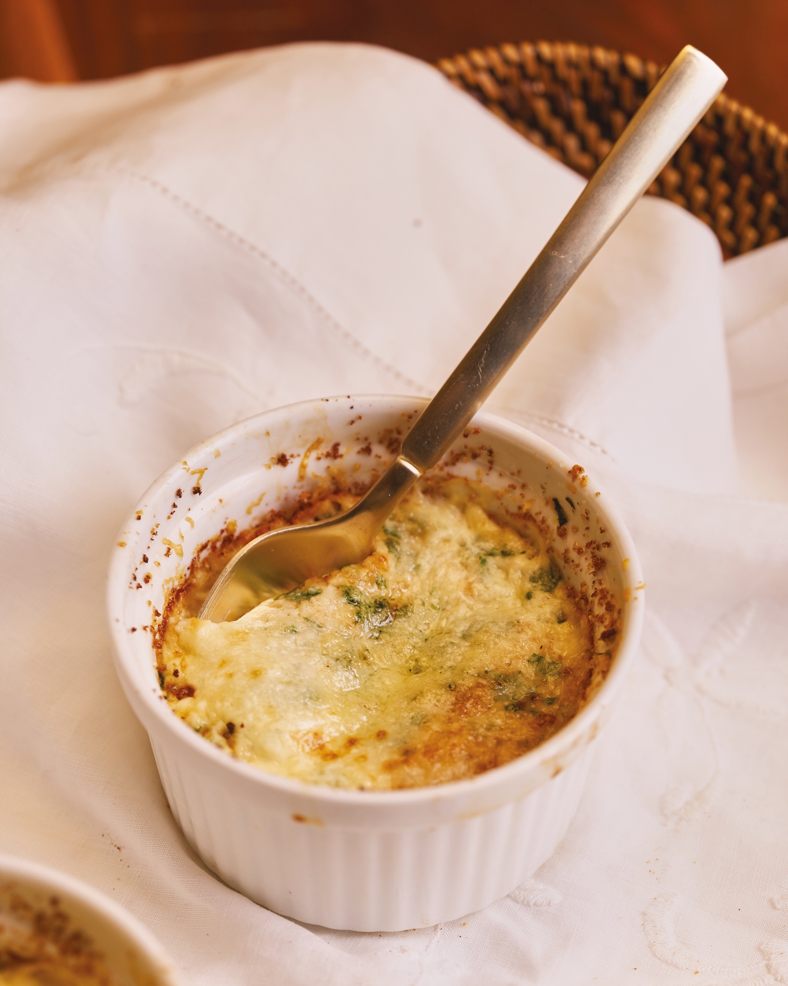 Close on one white ramekin of baked eggs, golden at the edges and flecked with herbs, a spoon lifting the first bite, on a white cloth in a basket.
