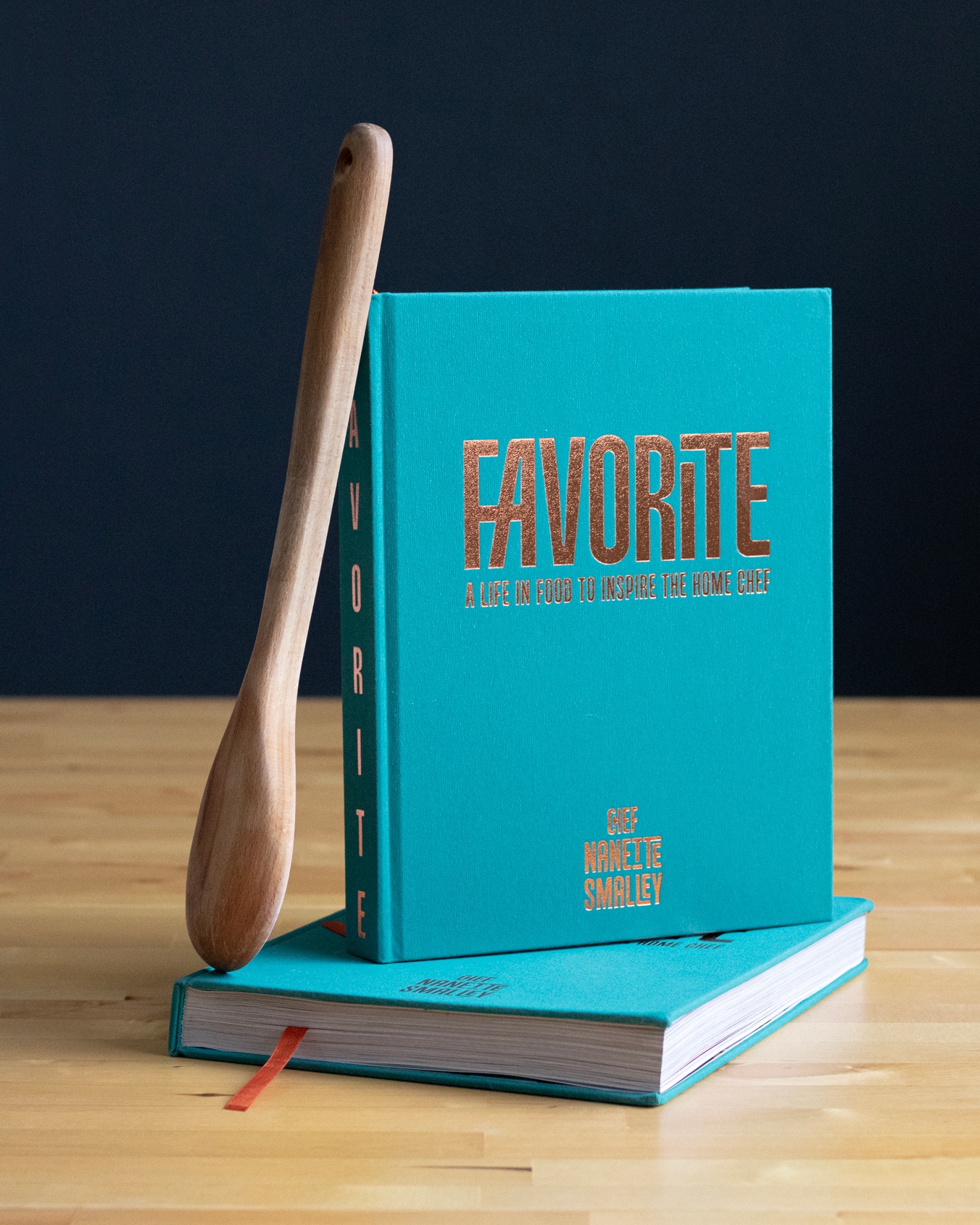 The book Favorite, in turquoise linen with copper foil lettering, standing on a second copy on a wooden table against a navy wall, a wooden spoon leaning against it and a copper ribbon marker showing.
