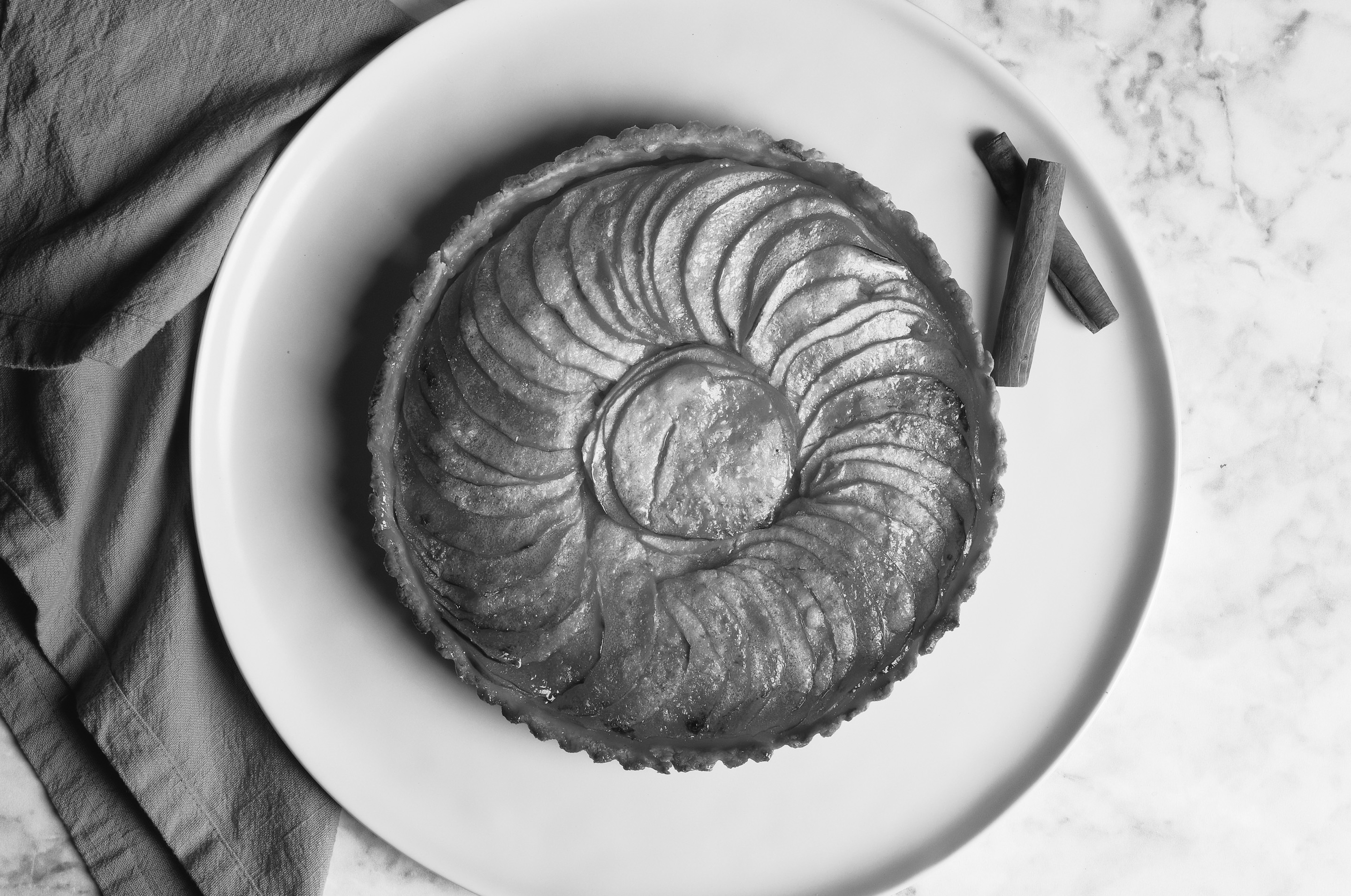 Black and white, seen from above: a French apple tart, its glazed apple slices laid in a spiral, on a pale plate on white marble, with a napkin at the left and two cinnamon sticks.