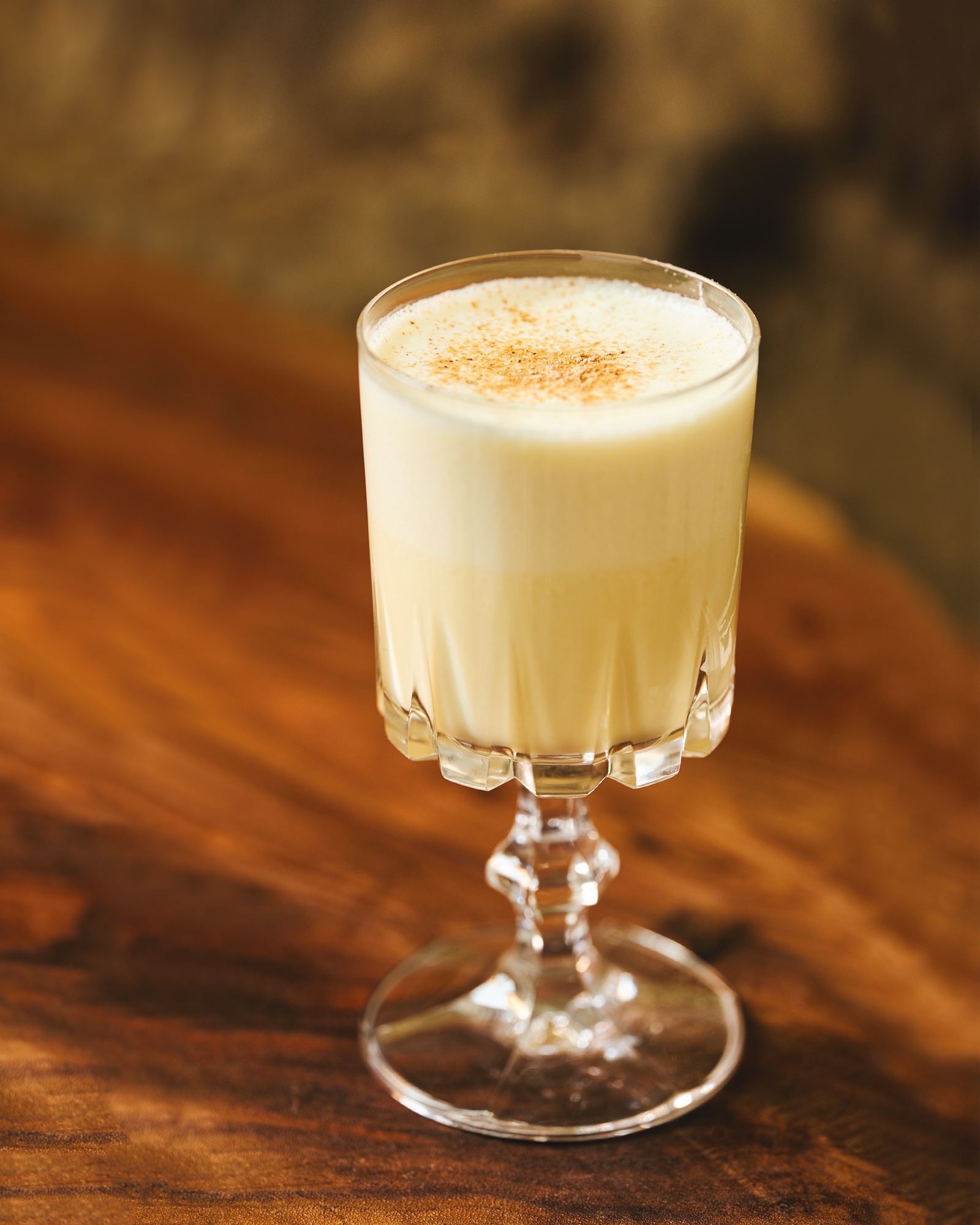 A cut-glass goblet of eggnog dusted with nutmeg, on a dark wooden table.