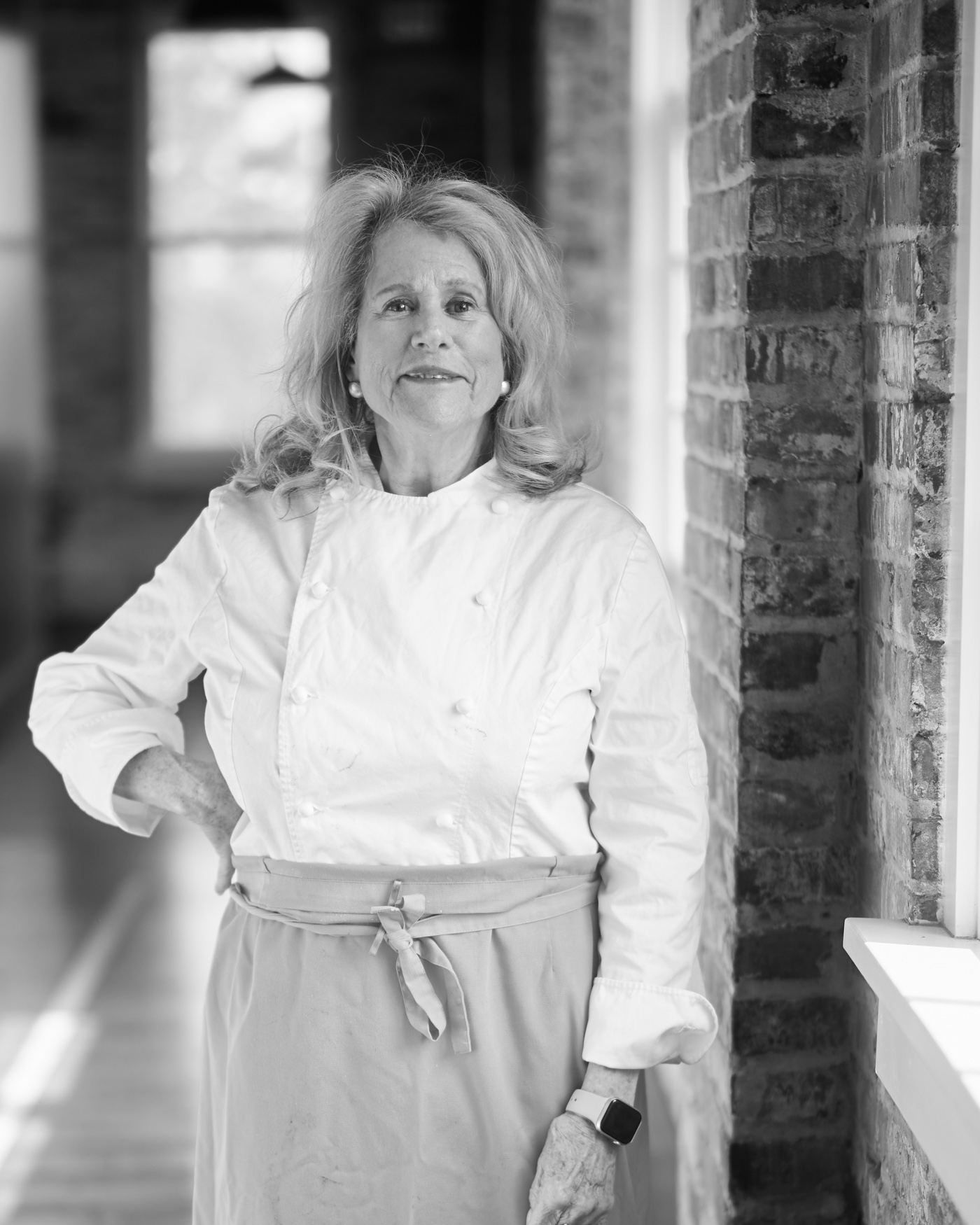Black and white: Nanette stands in a sunlit brick corridor in chef whites and a long apron, one hand on her hip, smiling at the reader.