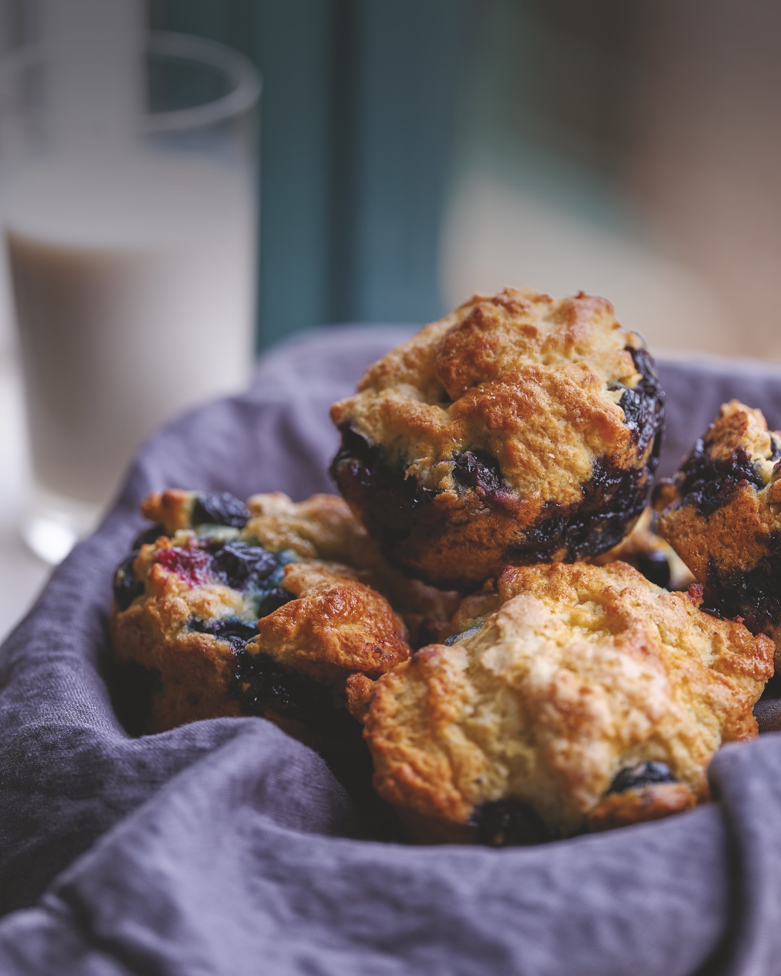 Blueberry muffins with golden, craggy tops and burst berries, piled in a basket lined with a grey-violet cloth, a glass of milk behind.