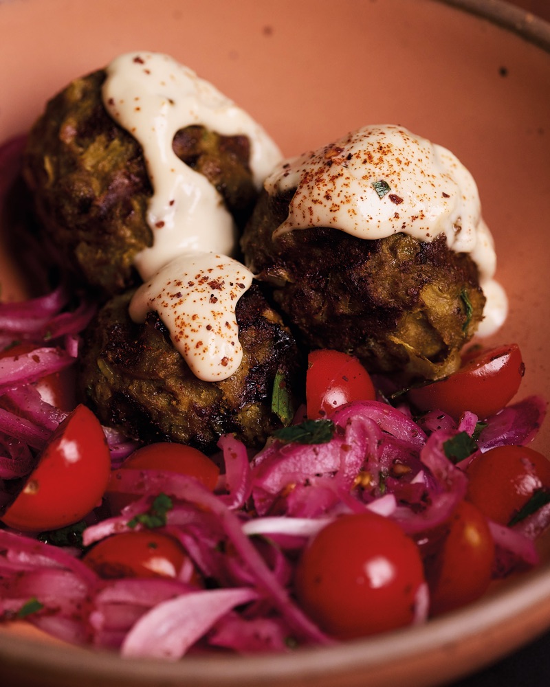 Three lamb meatballs under tahini sauce dusted with sumac, in a terracotta-pink bowl with halved cherry tomatoes and pink pickled onion.