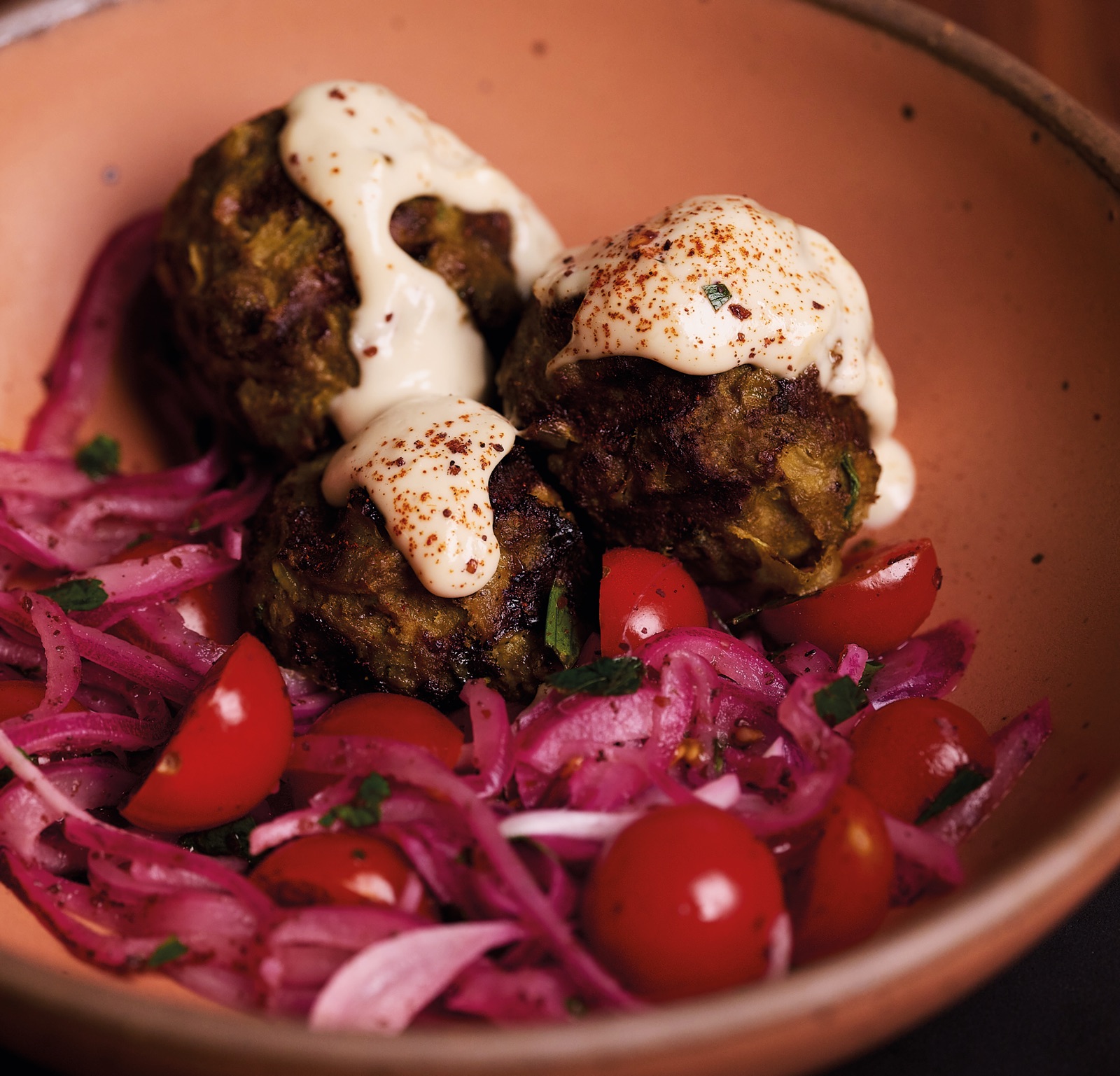 Three lamb meatballs under tahini sauce dusted with sumac, in a terracotta-pink bowl with halved cherry tomatoes and pink pickled onion.