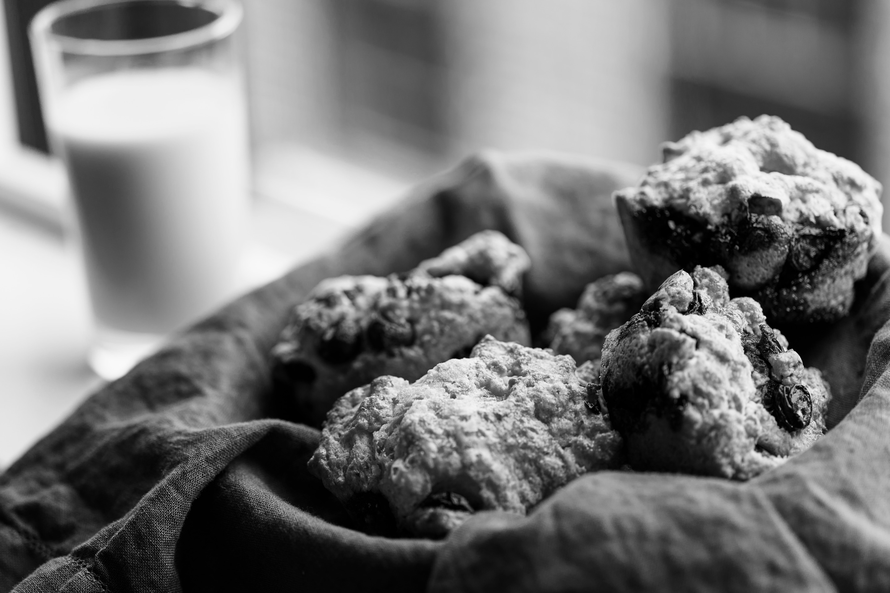 Black and white: blueberry muffins heaped in a linen-lined basket by a window, a glass of milk behind them.