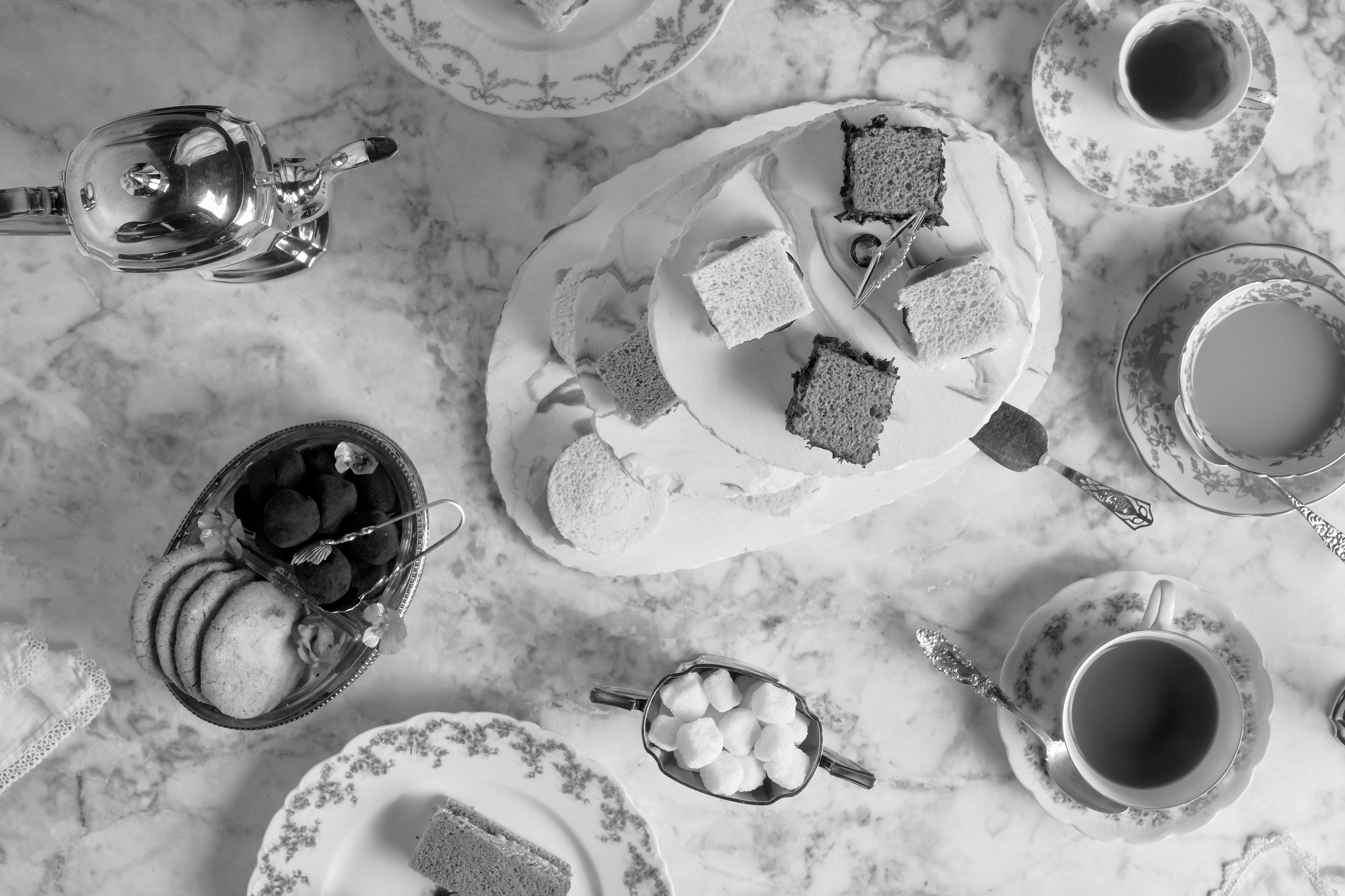 Black and white, from above: a tea table on marble, with a silver teapot, a tiered plate of crustless sandwiches, a dish of truffles and biscuits, sugar cubes, and cups of tea in rose-patterned china.