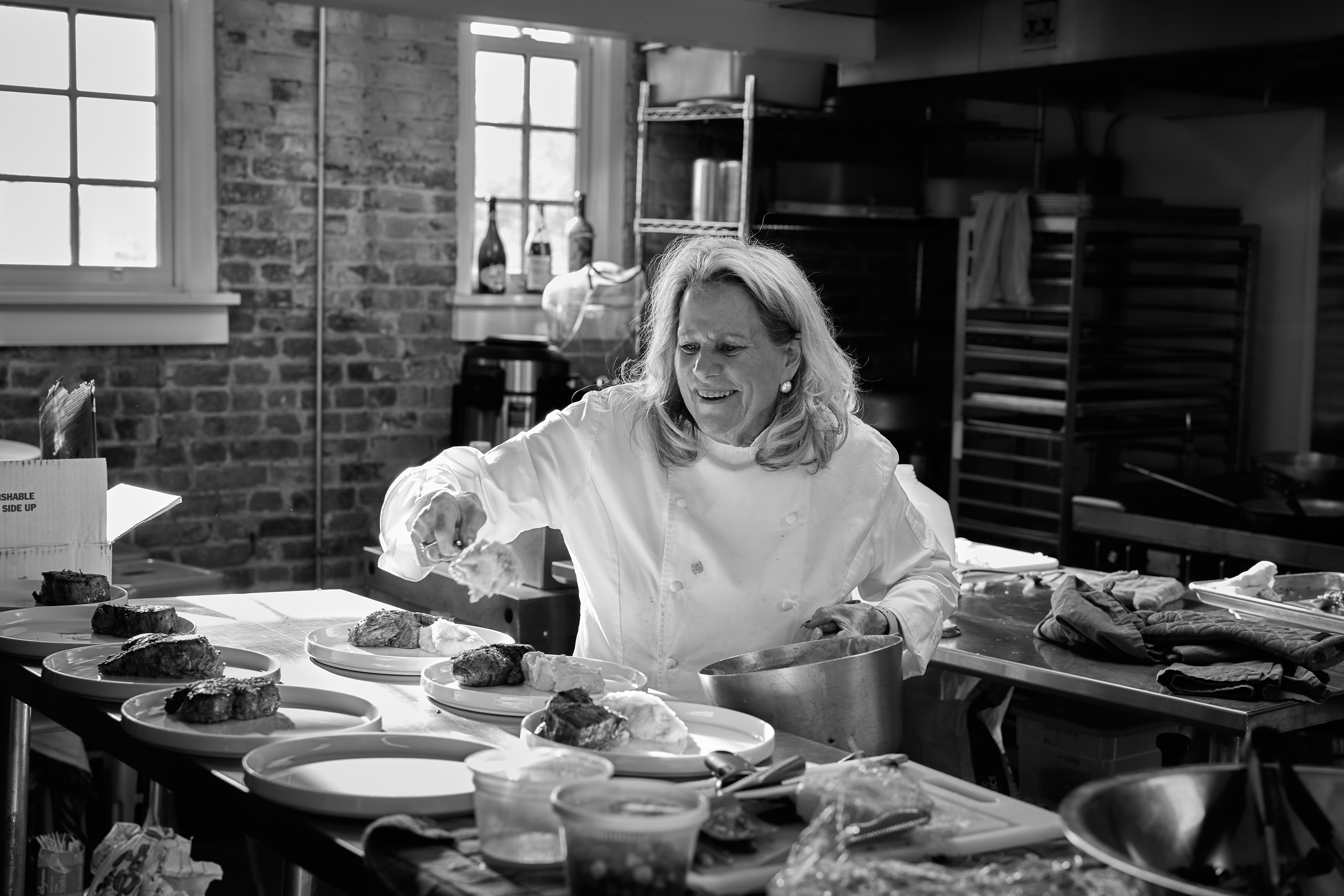 Black and white photograph: Nanette in her chef’s coat, smiling broadly, spoons potato onto a row of dinner plates in a brick-walled kitchen.