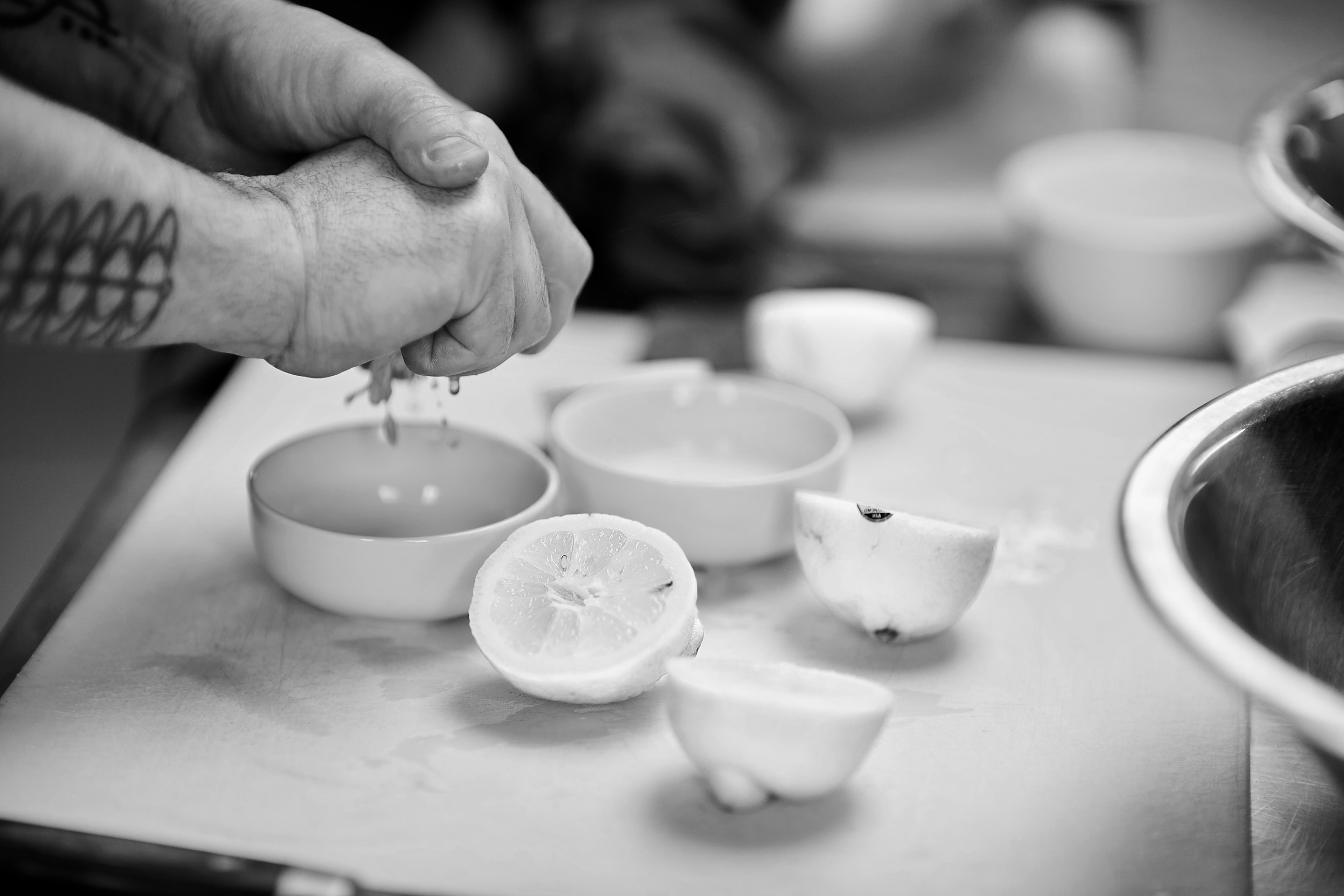 Black and white: a cook's hand squeezes half a lemon into a small white bowl; lemon halves and more bowls on a white board.
