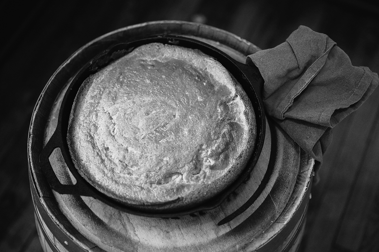 Black and white, from above: a round of cornbread, its top cracked and golden, in a cast iron skillet on a wooden barrel top, a napkin tucked at the handle.