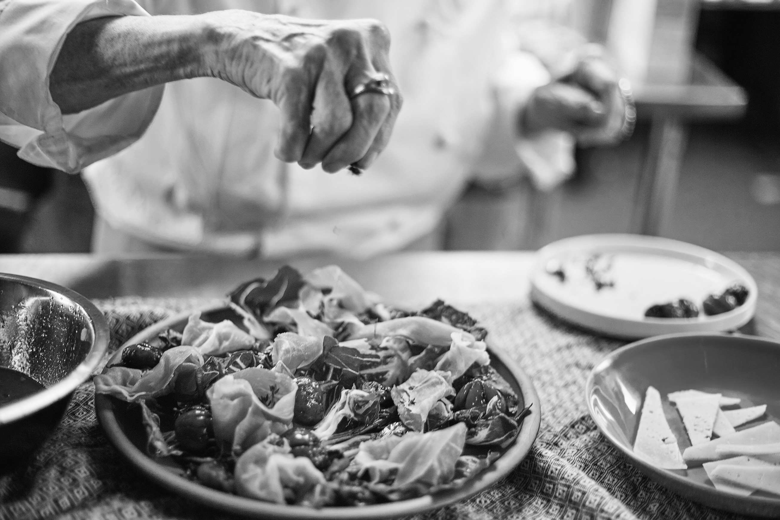 Black and white: Nanette’s hand, a gold ring on it, scatters herbs over a radicchio salad on a woven cloth, with a plate of sliced cheese beside it.