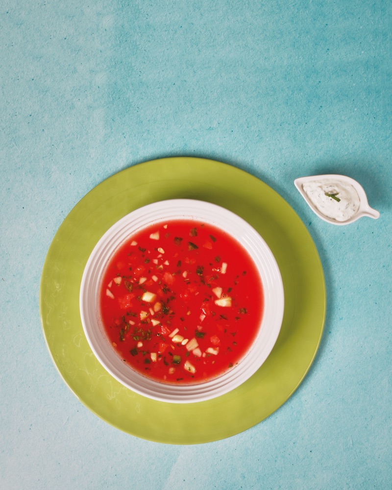 Seen from above on a turquoise ground: red watermelon gazpacho in a white bowl on a lime green plate, a small dish of herbed cream beside it.