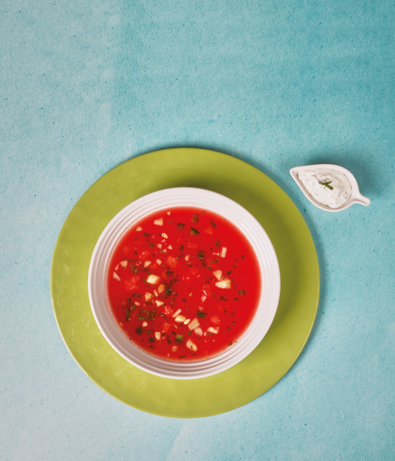 Seen from above on a turquoise ground: red watermelon gazpacho in a white bowl on a lime green plate, a small dish of herbed cream beside it.